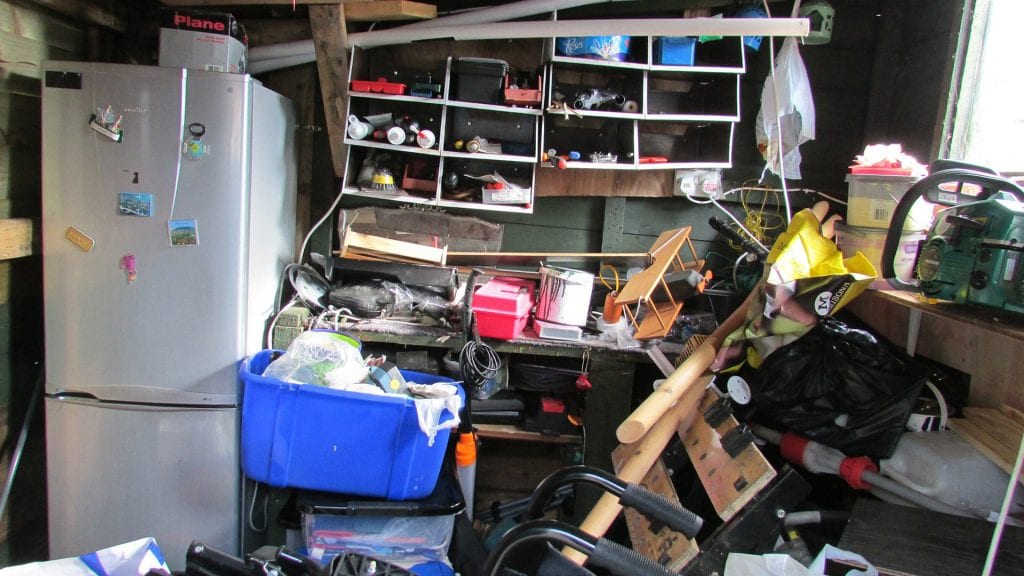 Cluttered storage area filled with household junk, including a refrigerator, blue bin, tools, and trash bags, representing typical items for Seattle Rubbish Removal services.
