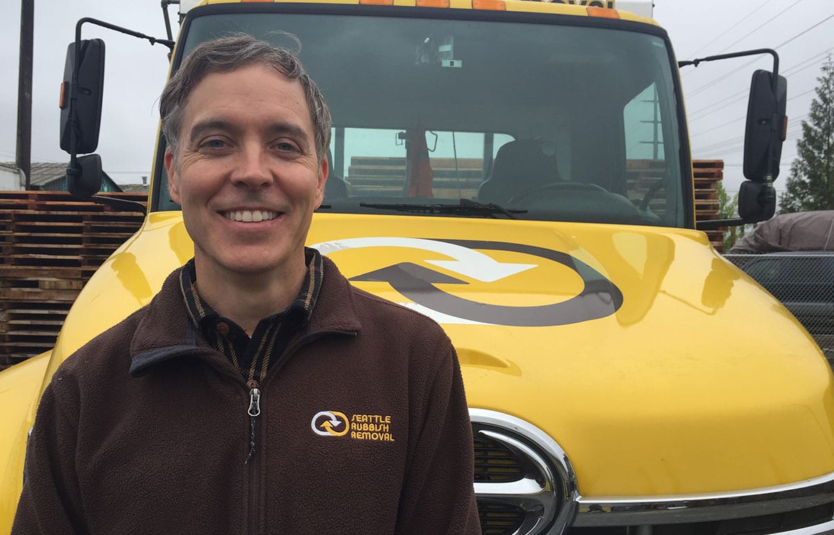 Owner of Seattle Rubbish Removal smiling in front of yellow junk removal truck, showcasing company branding and commitment to rubbish disposal services.