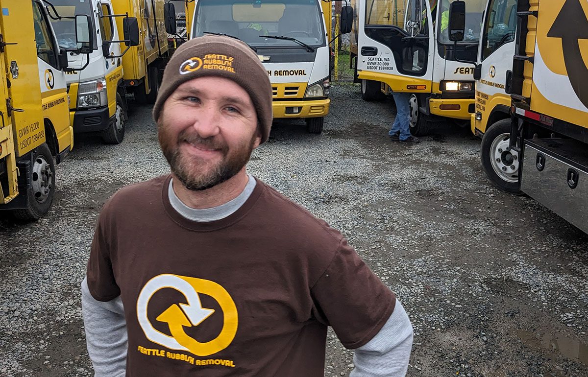 Crew member of Seattle Rubbish Removal smiling in front of company trucks, wearing a branded beanie and t-shirt, showcasing the team’s commitment to junk removal services.