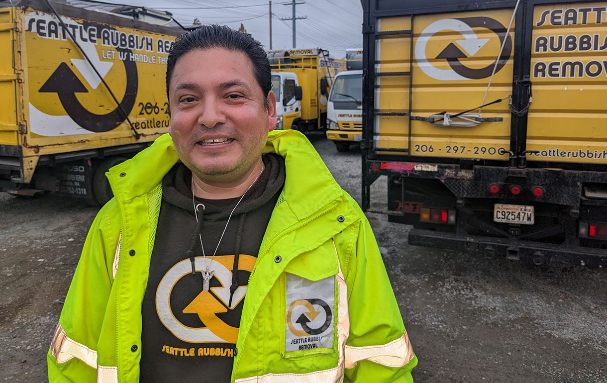 Omar from Seattle Rubbish Removal wearing a bright yellow safety jacket, smiling in front of company trucks, showcasing the team dedicated to junk removal services in Seattle.