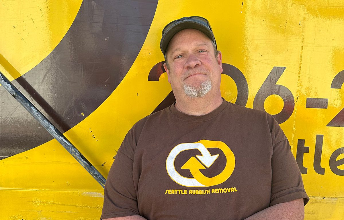 Man wearing a brown Seattle Rubbish Removal t-shirt standing in front of a yellow and brown logo background, representing the team's commitment to junk removal services in Seattle.