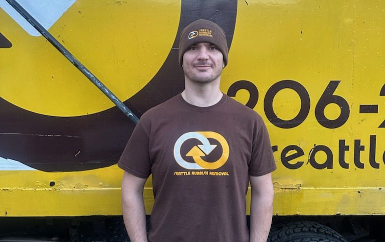Seattle Rubbish Removal crew member wearing company-branded shirt and beanie, standing in front of a yellow truck displaying the Seattle Rubbish Removal logo and contact information.