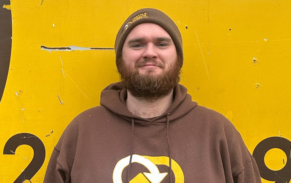 Seattle Rubbish Removal crew member wearing a brown hoodie and beanie, standing in front of a yellow background with a logo, representing the dedicated team behind junk removal services.